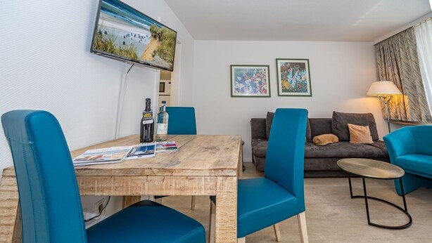 A modern living room with a wooden dining table, blue upholstered chairs, a grey sofa, and a small round table with a lamp. - Neptun - Wohnen am Meer, in Dieser Zentralen Wohnung (Sylt)