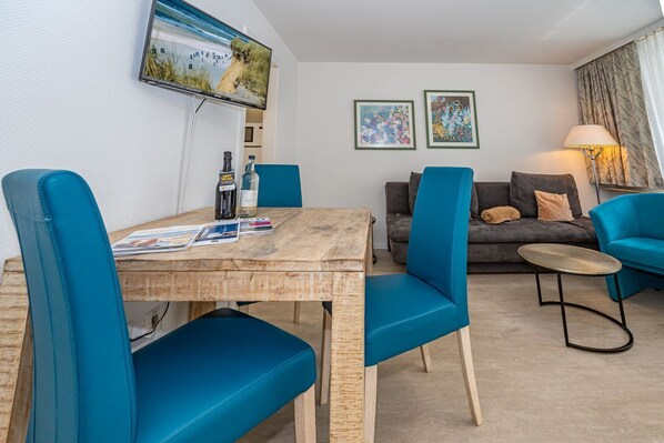 A modern living room with a wooden dining table, blue upholstered chairs, a grey sofa, and a small round table with a lamp.