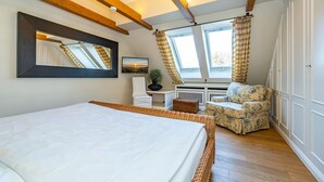Ein Schlafzimmer mit einem großen Bett, einem Sessel, einem kleinen Tisch und einem Fenster mit Vorhängen.