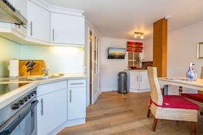 A compact kitchen with white cabinets, a stove, and a dining area with a wicker chair and table. - Üüs Lek - Neu!!! Ab Januar 2023 in Unserer Vermittlung. Sehr Gemütliche 2 Zimmerwohnung für max 4 (Sylt)