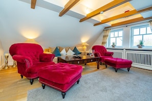 Un salon chaleureux avec un plafond haut et des poutres apparentes, comprenant un ensemble de canapés, une table basse et un pouf rouge.