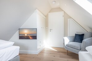 Une pièce moderne et bien éclairée, avec un plafond en pente, une grande fenêtre, un canapé gris et un tableau accroché au mur représentant un coucher de soleil sur l’océan.