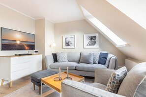 A modern living room with a grey sofa, a wooden coffee table, and a TV on a white cabinet. There are two framed artworks on the wall and a skylight above the sofa.
