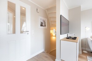 A modern, well-lit hallway with a white door, a framed artwork, a flat-screen TV mounted on a wall, and a desk with a lamp.
