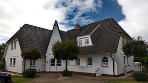 A two-story white house with a steeply pitched roof, a small front porch, and a paved driveway. - Witt Hingst App. 10 - Cozy 2-room vacation apartment, quietly situated with (Sylt)