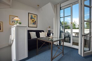Een kamer met een witte koelkast, een tafel met glazen blad, een donkere bank, een lijst met schilderij en een groot raam met uitzicht op de omgeving.