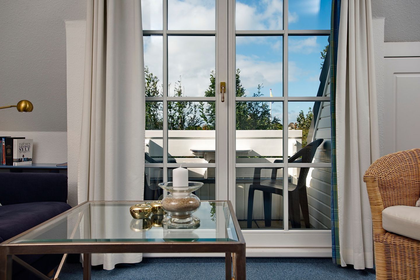 Ett modernt vardagsrum med ett soffbord i glas, en blå soffa, en fåtölj i rotting och utsikt över utomhusmiljön genom en skjutdörr i glas.