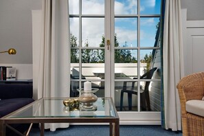 Ein modernes Wohnzimmer mit einem Glastisch, einem blauen Sofa, einem Korbstuhl und Blick ins Freie durch eine Schiebetür.