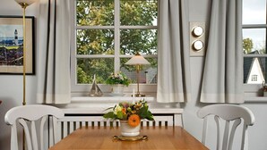 A dining area with a wooden table, white chairs, a window with curtains, a flower vase, and a lamp. - Witt Hingst App. 10 - Cozy 2-room vacation apartment, quietly situated with (Sylt)