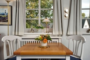 Un coin repas avec une table en bois, des chaises blanches, une fenêtre avec des rideaux, un vase de fleurs et une lampe.