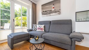 Una sala de estar moderna con un sofá gris, un reposapiés a juego y una mesa de centro hexagonal. Un cuadro de un faro adorna la pared, y hay una ventana con cortinas.