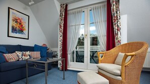 Un salon comprenant un canapé, un pouf et un fauteuil en osier, une table en verre et un tableau accroché au mur.
