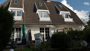 Une maison à deux étages, dotée d’un toit en pente, de lucarnes et d’un porche à l’avant, aménagé avec des sièges pour l’extérieur.