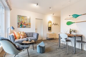Una sala de estar moderna con un sofá gris, una mesa de comedor con sillas, una pequeña zona de cocina y un cuadro abstracto colorido en la pared.
