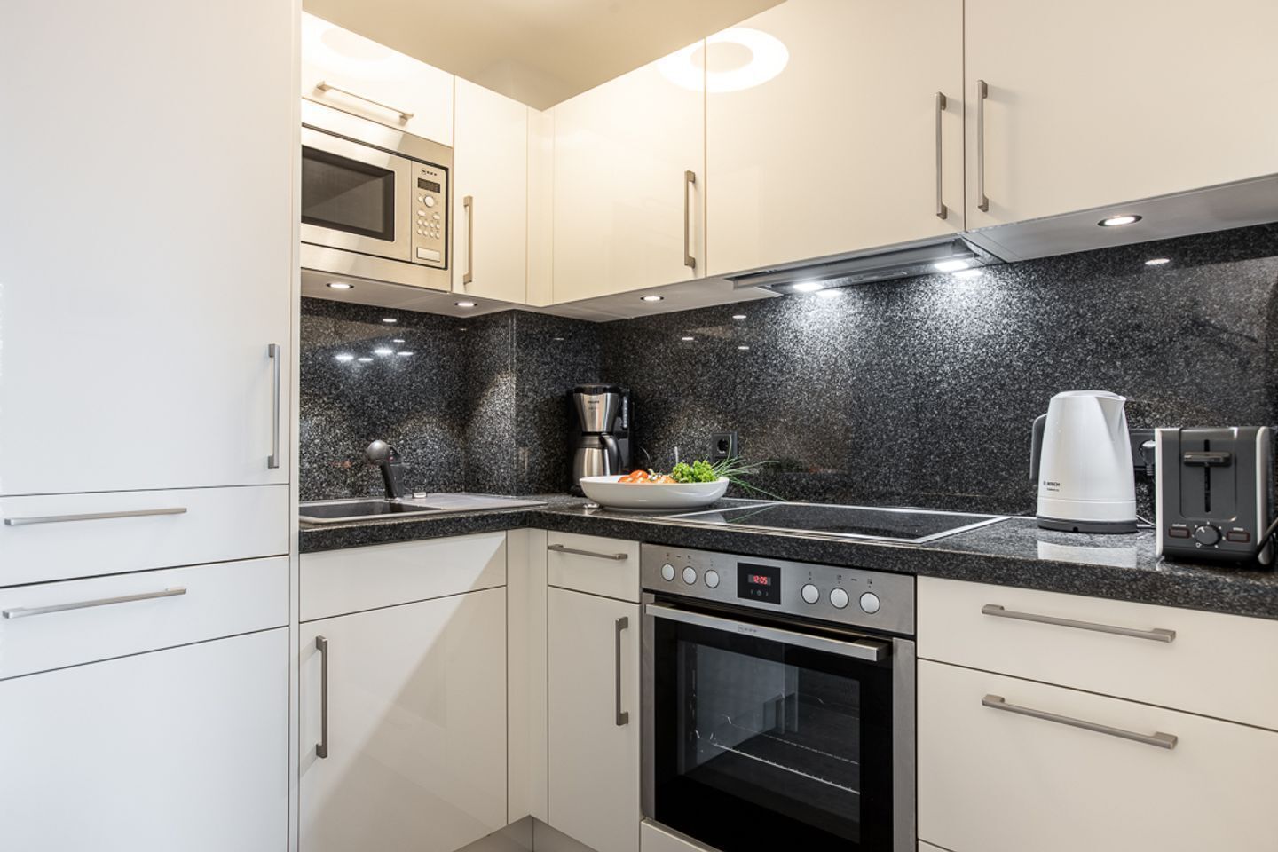 A modern kitchen with white cabinets, a black granite countertop, a microwave, a coffee maker, a toaster, and an oven.