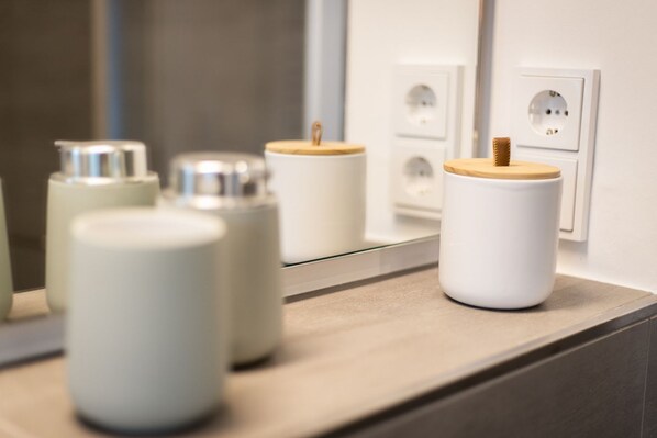 A bathroom counter with four white containers with wooden lids, a mirror reflecting the containers and a wall socket.