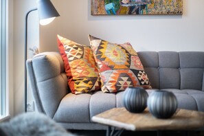 Een grijze bank met twee gekleurde sierkussens, een zwarte vloerlamp en een klein houten bijzettafeltje met twee decoratieve vazen.