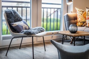 A modern living room with a grey chair with a furry throw, a small wooden table with a vase, and a sofa with patterned cushions.