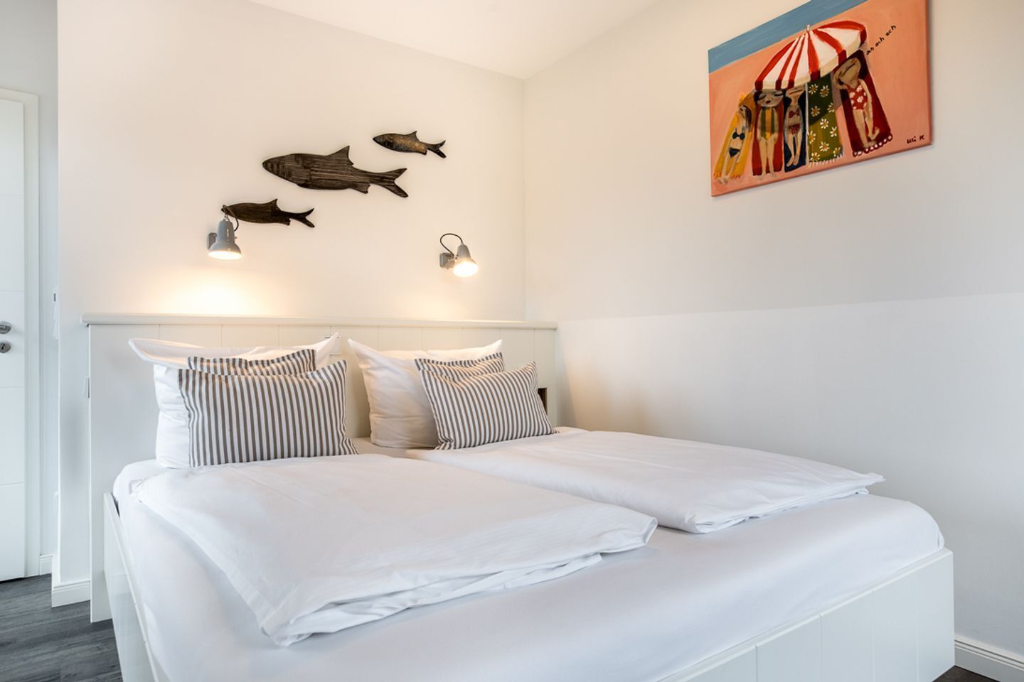 Une chambre d’hôtel avec un lit, des oreillers à rayures, des lampes fixées au mur et un tableau représentant une scène de plage.