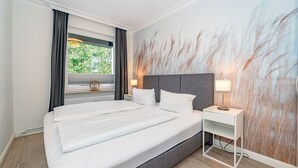 Ein Hotelzimmer mit einem großen Bett, einem Nachttisch mit Lampe, einem Fenster mit Vorhängen und einer Wand mit einem strukturierten Wandgemälde.