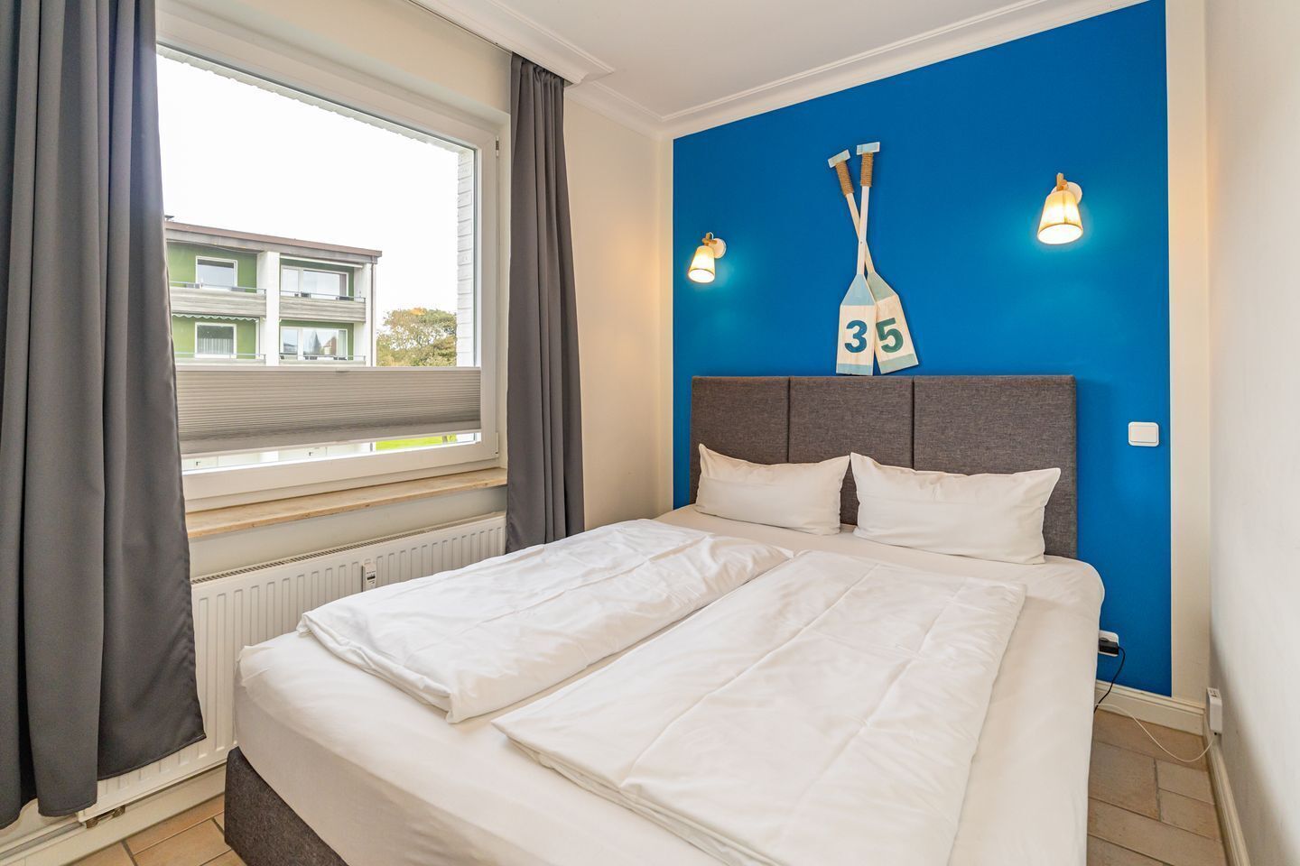 A bedroom with a bed, a window with curtains, a blue wall with a number sign, and two wall-mounted lights.