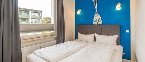 A bedroom with a bed, a window with curtains, a blue wall with a number sign, and two wall-mounted lights.