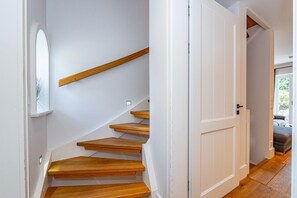 Eine Treppe mit Holzdielen führt zu einer weißen Tür, ein kleines Fenster mit einer Pflanze und ein Blick auf ein Bett im angrenzenden Zimmer.
