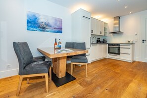 Moderni keittiö, jossa on valkoiset kaapit, sisäänrakennettu uuni sekä ruokailutila, jossa on puinen pöytä ja kaksi tuolia.