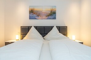 A neatly made bed with white linens and two bedside lamps, a headboard with a textured design, and a painting of a beach scene above the bed.