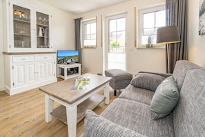 A living room with a sofa, a coffee table, a TV, and a cabinet with glass doors. - Relax App.2 - Beautiful apartment in the south of Westerland with terrace. (Sylt)