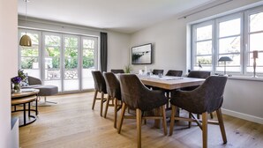 A dining area with a wooden table and chairs, large windows with curtains, and a view of greenery outside.