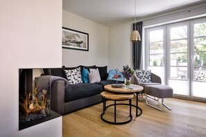 Ein modernes Wohnzimmer mit einem dunkelgrauen Sofa, einem runden Couchtisch aus Holz und einem grauen Sessel. Ein gerahmtes Bild ist an der Wand aufgehängt, und ein Fenster bietet einen Blick ins Freie.