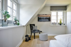 Ein Schlafzimmer mit schrägem Dach, einem Bett, einem Stuhl, einem Gemälde und einem Fenster mit Pflanzen.