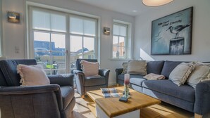 Una sala de estar moderna con un sofá, sillones y una mesa de centro. Amplias ventanas ofrecen vistas al exterior.