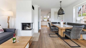 Un salon moderne avec une cheminée, un coin repas avec une table et des chaises en bois, et une cuisine en arrière-plan.