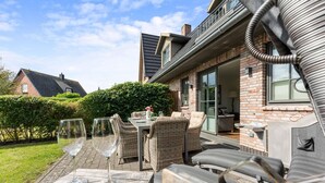 Une terrasse extérieure avec des meubles en osier et des verres à vin, une maison en briques avec une porte en verre, et une haie en arrière-plan.