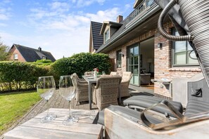 Une terrasse extérieure avec des meubles en osier et des verres à vin, une maison en briques avec une porte en verre, et une haie en arrière-plan.