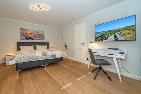 A bedroom with a bed, a desk, a chair, a TV, and a painting on the wall.