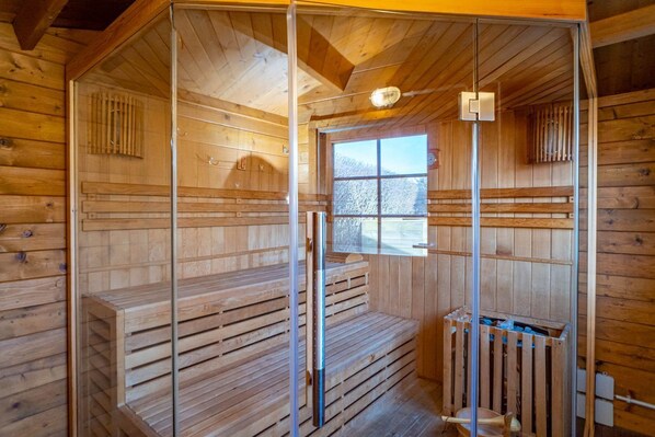 Una sauna de madera con un panel de vidrio, un banco y una ventana con vistas a árboles cubiertos de nieve.