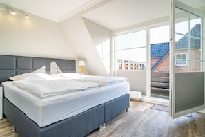 A modern bedroom with a large bed, a grey headboard, and a view of a brick building through a sliding glass door. - Haus Strandoase - NEU NEU Neu!!! Ab Januar 2023 bei uns in der Vermittlung. - Mit Süd Terrasse und (Sylt)