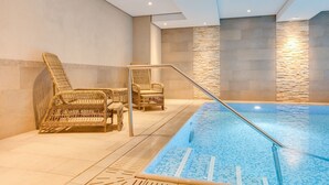 Una piscina interior con suelo de baldosas, rodeada de sillones de mimbre y una pared de piedra.