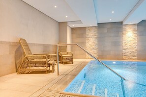 An indoor swimming pool with a tiled floor, surrounded by wicker chairs and a stone wall.