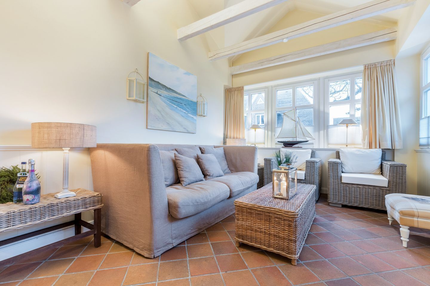 Ein gemütliches Wohnzimmer mit einem grauen Sofa, einem Korb-Couchtisch und einem Korbstuhl. An der Wand hängt ein Gemälde und es gibt ein Fenster mit Vorhängen.