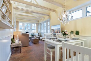 A spacious dining area with a white table set for four, a chandelier, and a view of the sea through large windows. - App. Wellness - Zentrale Stadtwohnung für 4 Personen mit Terrasse und Wellnessbereich (Sylt)