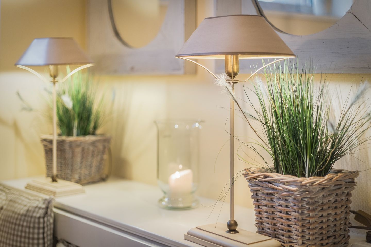 A room with a bedside table, a lamp, a candle, and a woven basket with a plant.