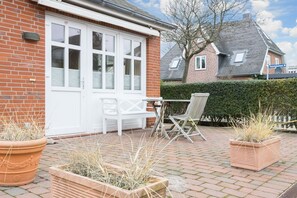 Une maison en briques avec une porte et des fenêtres blanches, une terrasse avec une table et des chaises, et deux plantes en pot.