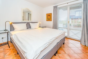 A neatly made bed in a hotel room with a large window, a bedside table with a lamp, and a framed picture on the wall. - Wildente App. 5 - Zentral Gelegene 2-zimmerwohnung in Wenningstedt am Dorfteich (Wenningstedt-Braderup (Sylt))