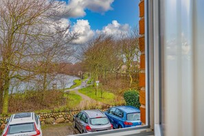 Blick auf einen Parkplatz mit zwei Autos, einem Weg, der zu einem See führt, und Bäumen im Hintergrund.