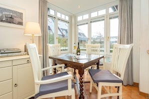 A dining area with a wooden table, white chairs, a lamp, and a bottle of wine. - Wildente App. 5 - Zentral Gelegene 2-zimmerwohnung in Wenningstedt am Dorfteich (Wenningstedt-Braderup (Sylt))