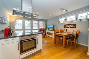 Una cocina moderna con horno empotrado, un hervidor rojo y un comedor con sillas de madera y una mesa.